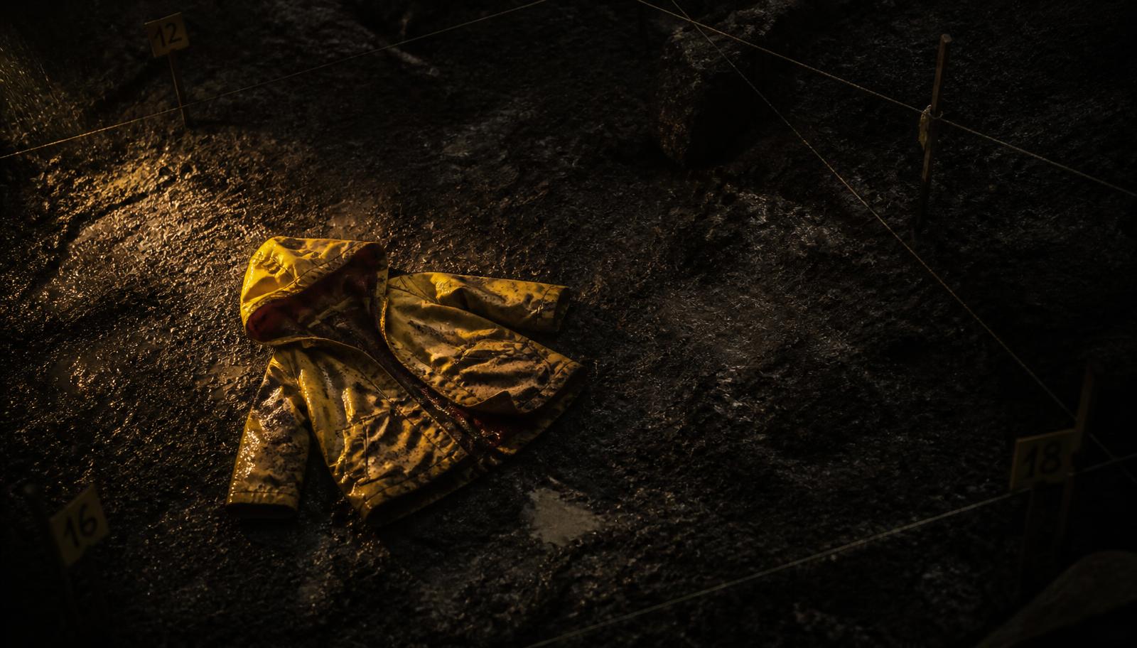 Original illustration: an empty child's raincoat lying on wet excavated earth inside a forensic survey grid, lit by a single low work-lamp